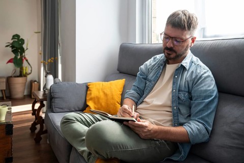 A man with glasses sits on a comfortable sofa, writing in a notebook with a pen. Sunlight streams