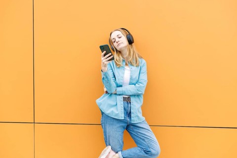 A stylish woman leans against an orange wall in Warsaw, wearing headphones and checking her phone,