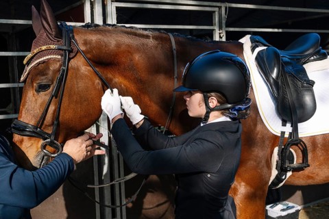 A teenager in classical dressage attire adjusts a horse's bridle with assistance, focusing intently