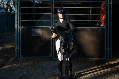 A teenage girl in classical dressage attire confidently holds a saddle. She stands by a stable,