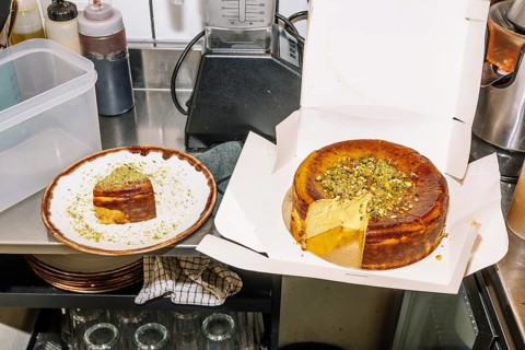 A rich cheesecake topped with crushed pistachios sits in an open box, with a slice served on a