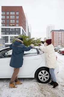 A couple wearing winter clothing secures a Christmas fir onto their car's roof in a snow-covered