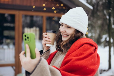 A cheerful young woman in a white beanie and red scarf smiles while holding a coffee cup and taking