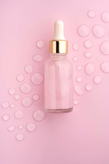 A clear glass dropper bottle on a pink background, surrounded by scattered water drops The scene