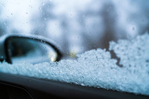 Close-up of a frosted car window with a blurred side mirror visible. The ice crystals and droplets