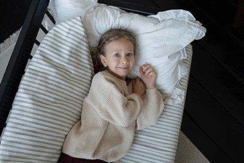 A child with light hair and blue eyes lies comfortably in bed, wearing a cozy sweater The bed is