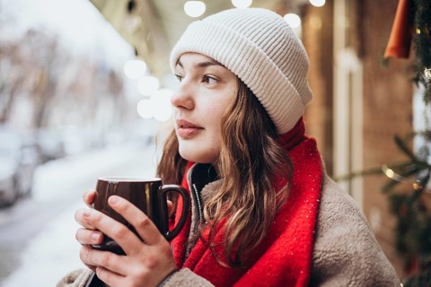 A young woman sips a hot chocolate on a snowy winter day. Dressed warmly in a white beanie and red