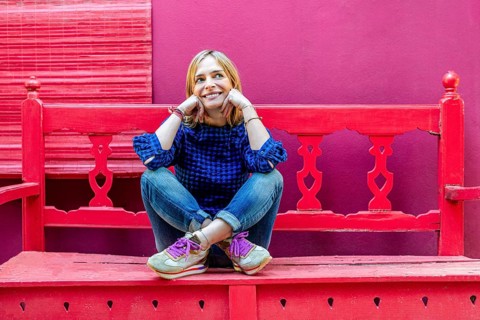 A woman wearing a blue plaid shirt sits cross-legged on red wooden stairs, smiling gently and