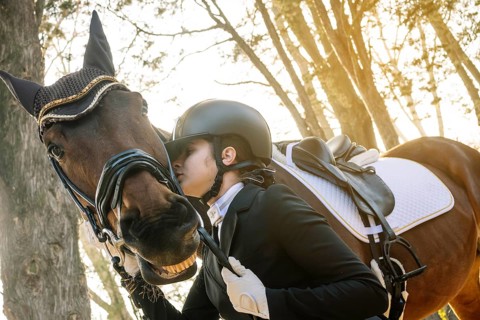 A teenage girl in classical dressage attire gently embraces her horse in a peaceful forest setting,