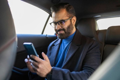 Indian man in blue turtleneck and black coat, using smartphone in car, looking at mobile screen