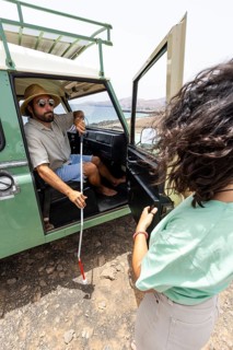 A blind man holds his cane as he steps out of a motorhome, with a woman nearby offering support.