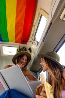 Lesbian couple laughing inside a van decorated with a rainbow flag. One holds a book while the