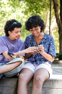 A cheerful multiethnic LGBTQ+ couple sitting outdoors, sharing and looking at a phone together,