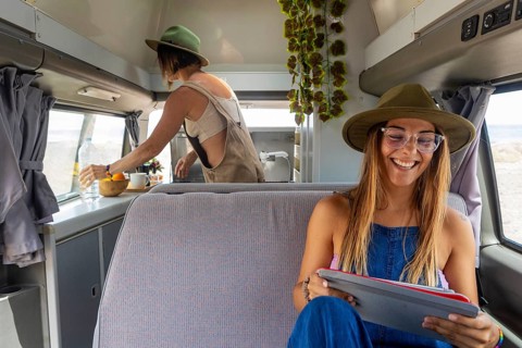 A lesbian couple relax inside a camper. One prepares a meal at the counter while the other enjoys
