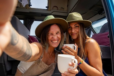 Lesbian couple wearing hats enjoy coffee inside a camper van, capturing a joyful moment of