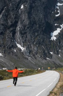 A traveler in a vibrant jacket walks along a winding road, surrounded by Norway's majestic