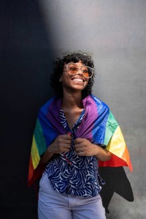 A smiling LGTBQ+ person wearing sunglasses and a patterned shirt, joyfully wrapped in a rainbow