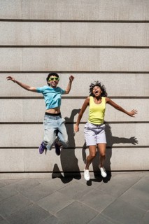 A joyful, multiethnic LGBTQ+ couple, wearing vibrant summer attire, jumps against a concrete wall,