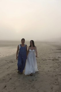 Two women walk along a foggy beach, one in overalls and the other in a flowing white dress The