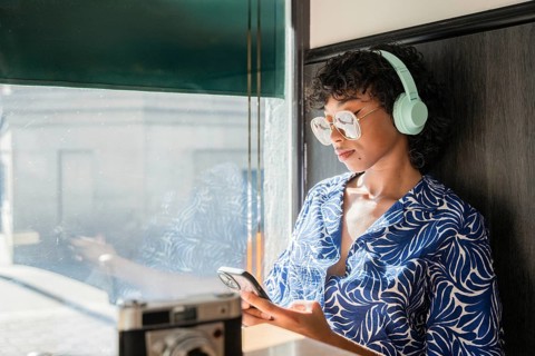 A stylish young woman with curly hair and glasses wearing headphones, leans against a window while