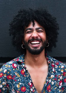 A man with curly hair laughs joyfully, wearing a vibrant floral shirt against a gray background.