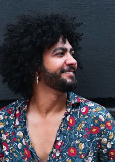 A man with curly hair and a beard smiles while wearing a vibrant floral shirt against a gray