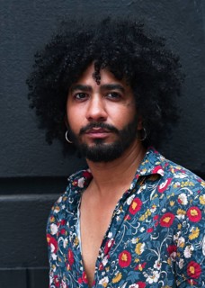 A close up portrait of a man with curly hair, wearing a floral shirt and hoop earrings against a