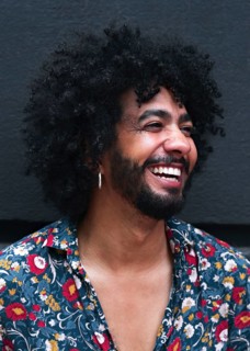 A smiling man with curly hair and a floral shirt exudes happiness. Their joyful expression and