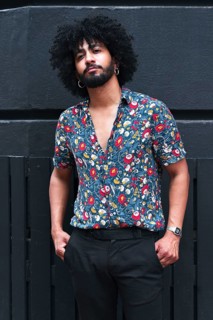 A stylish individual with curly hair and beard leans against a gray wall, wearing a vibrant floral