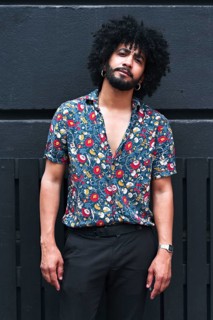 A confident young man with curly hair poses in a vibrant floral shirt against a gray urban wall,