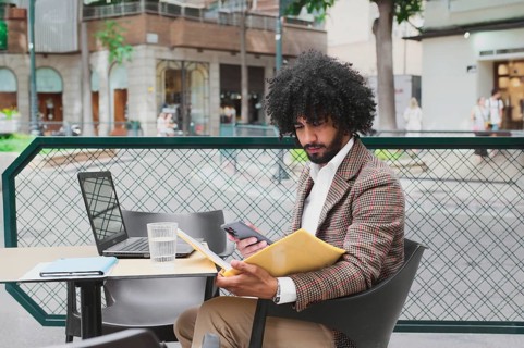 A Mixed-race man with curly hair is engaged in remote work using a laptop and smartphone at an