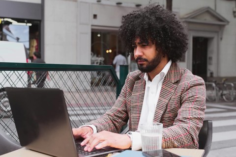 A mixed-race male with curly hair is teleworking at an outdoor cafe in a bustling city environment,