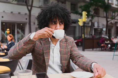 A Mixed-race man with curly hair teleworks from a street cafe, sipping coffee, dressed in a stylish