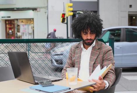 A young Mixed-race man with curly hair working remotely at a street-side cafe, carefully reviewing