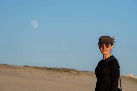 A smiling woman dressed in casual clothes and sunglasses stands on a sandy beach looking at camera