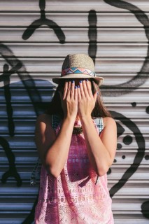 A woman in a straw hat and pink dress stands against a graffiti wall, covering her face with her