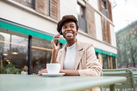 Black woman sits at an outdoor cafe, smiling and talking on her smartphone She is wearing a stylish
