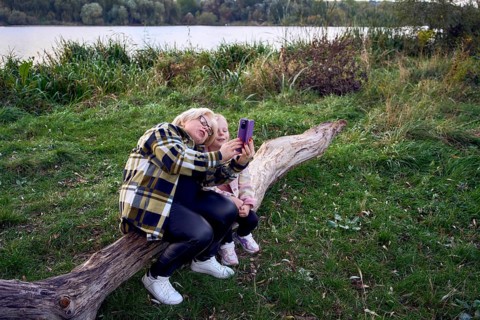 A mother and daughter sit on a wooden log by a scenic riverside, capturing a joyful selfie together