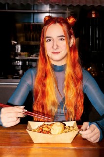A woman with vibrant red hair sits at an Asian food market, savoring a meal with chopsticks The