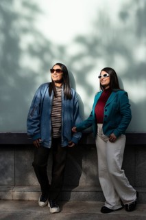 A stylish Latin lesbian couple enjoys a sunny day, dressed in casual fashion with jackets and