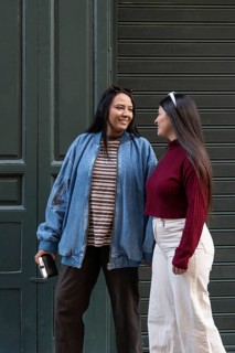 A Latin lesbian couple shares a joyful moment outdoors. One wears a denim jacket and striped shirt,