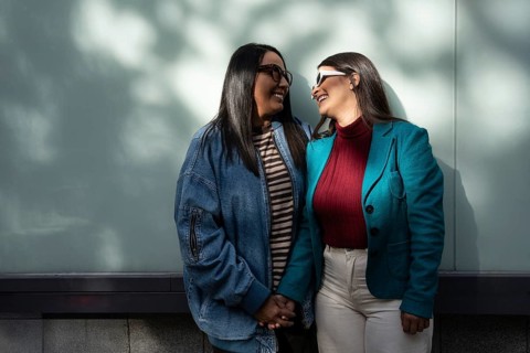 A Latin lesbian couple stands closely, smiling at each other with affection. They hold hands,