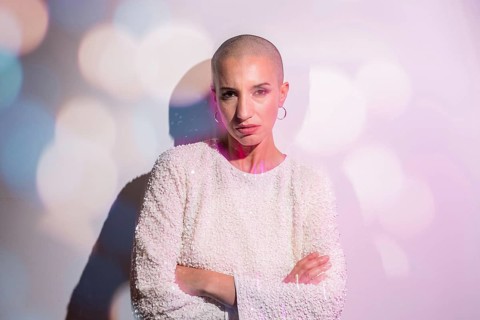A middle-aged woman with a shaved head stands confidently against a softly lit backdrop, embodying