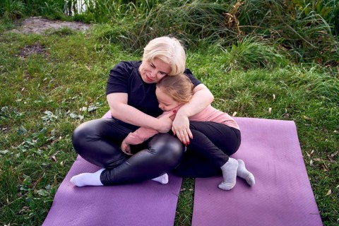 A mother and his daughter share a warm, comforting hug while sitting on yoga mats in a riverside.