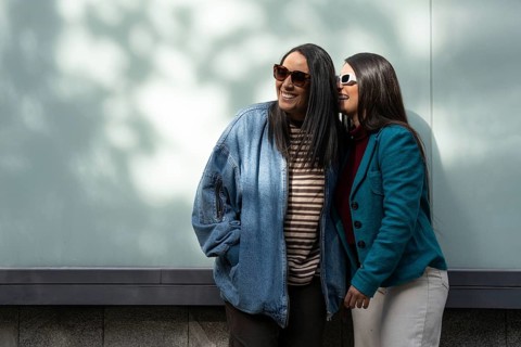 A Latin lesbian couple stands close, basking in the sun. One wears a denim jacket, the other a teal