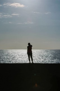 Silhouette of a person standing by the sea during sunset. The calm water and warm light create a