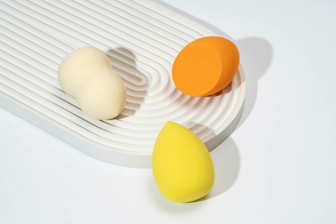 A trio of colorful cosmetic sponges in orange, yellow, and cream are arranged on a textured, white