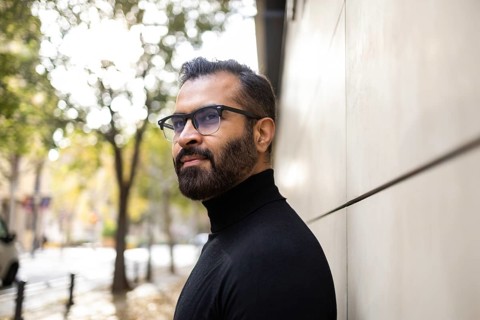 Side view of an Indian man dressed in a black turtleneck stands thoughtfully on a city street lined