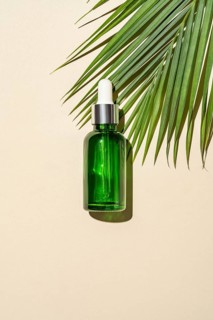 Top view of a green glass serum bottle with a dropper pipette, cast by a palm leaf's on a warm