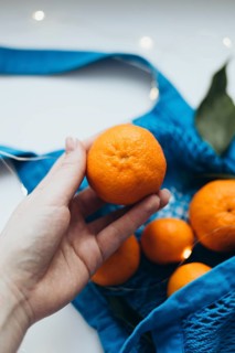 Cropped unrecognizable image of a hand holding a tangerine over a blue string bag filled with